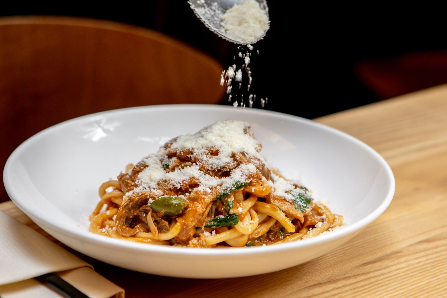 A hand sprinkles grated cheese over a plate of pasta with tomato sauce and vegetables, served in a white bowl on a wooden table.