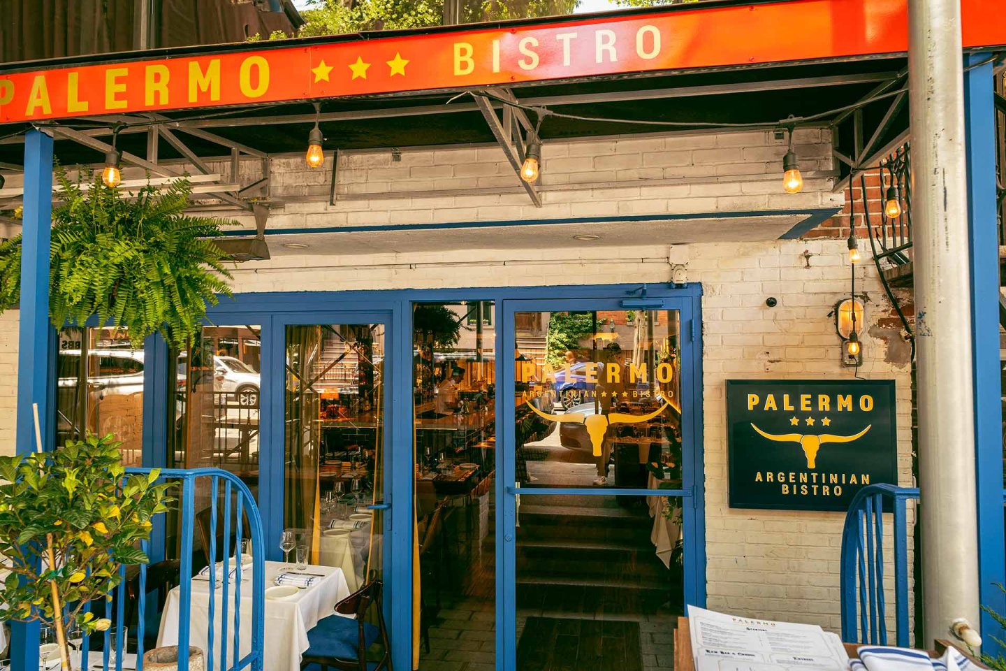 Exterior of Palermo Argentinian Bistro with a glass door, blue window frames, hanging lights, a green plant, and a yellow sign displaying the restaurant name on a white brick wall. Tables are set on the patio.