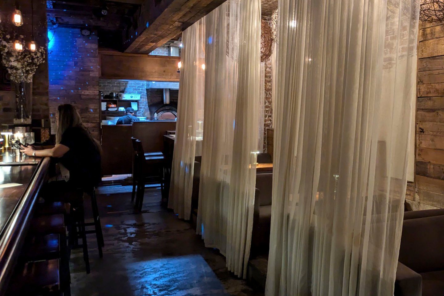 A dimly lit restaurant interior with sheer curtains partially sectioning off seating areas. A person sits at a bar on the left, and there are wooden accents throughout the space. Subtle blue lighting adds ambiance.