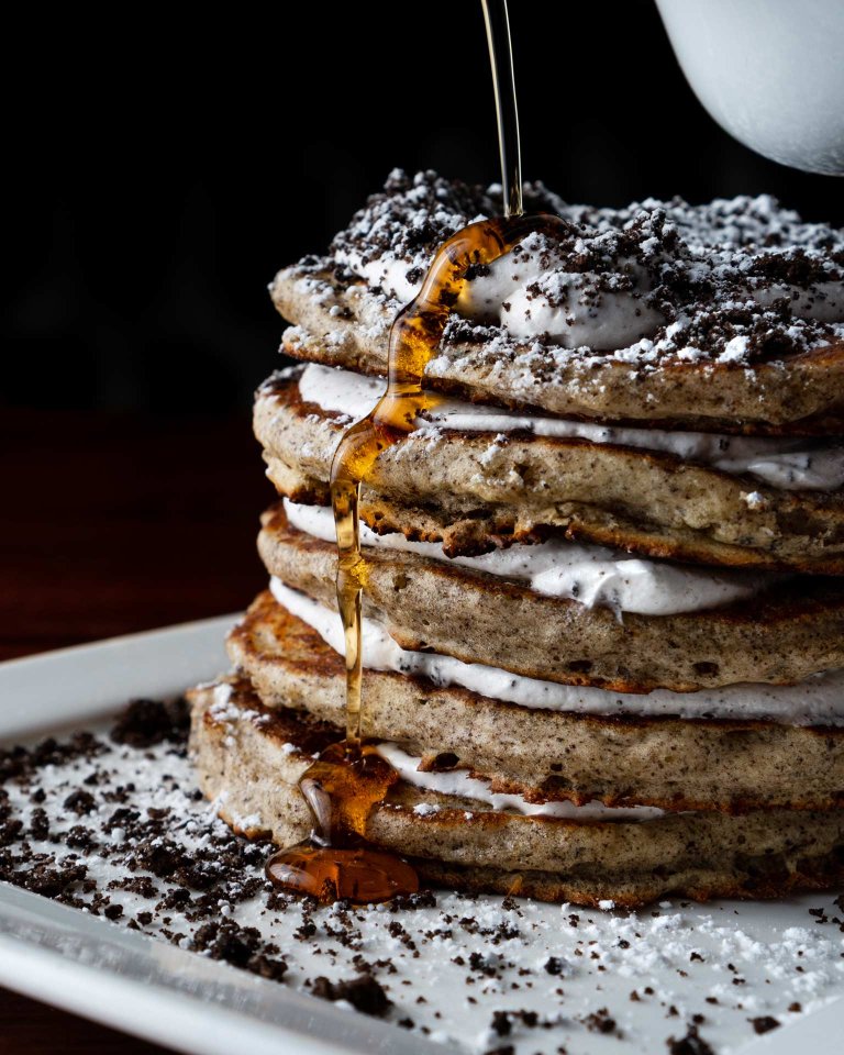A stack of pancakes layered with cream and cookie crumbles is shown on a white plate, with syrup being poured over the top, creating a glossy drizzle down the side.