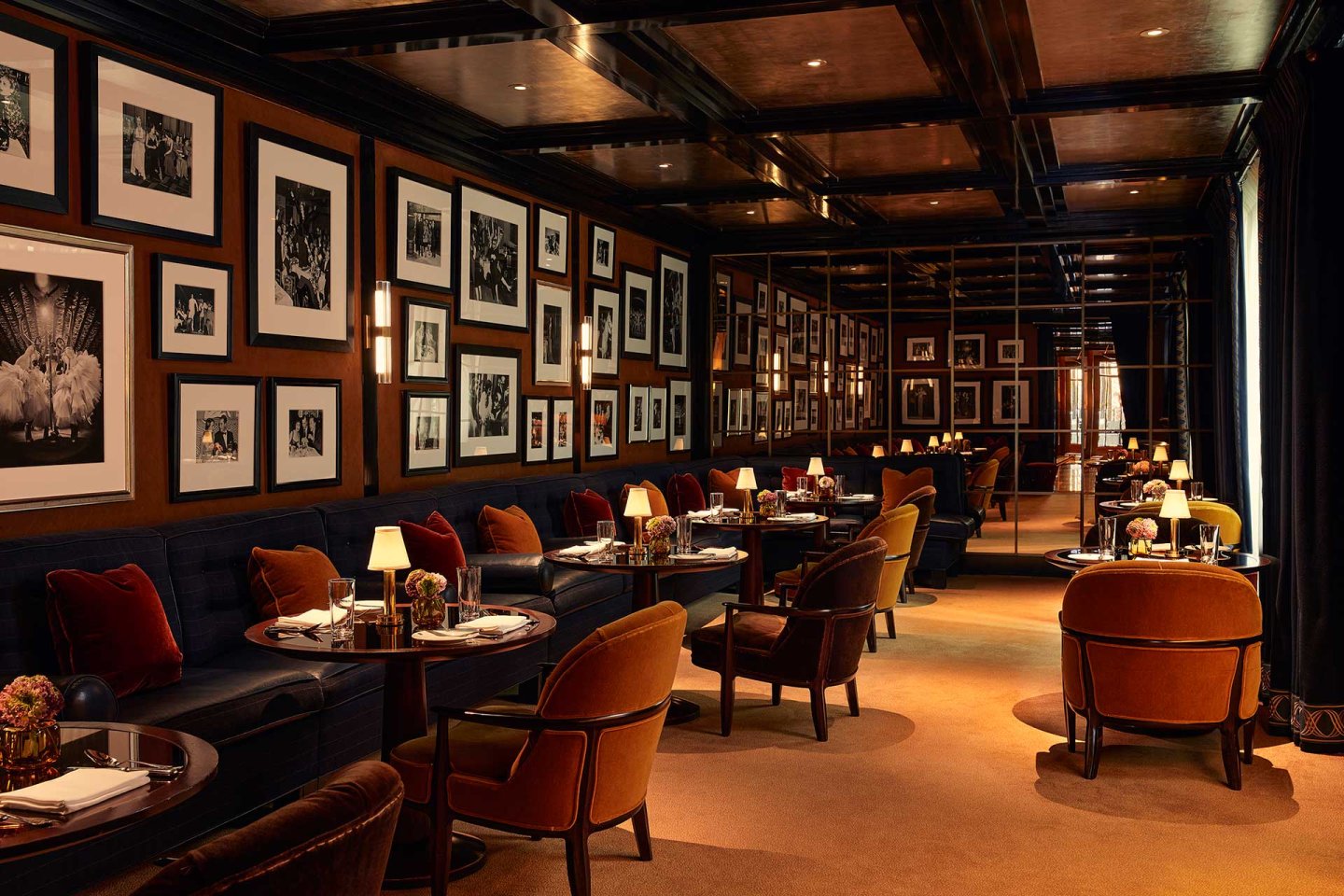 Elegant restaurant interior with dark wood paneling, framed black and white photos on the walls, plush velvet chairs, small round tables set for dining, and warm ambient lighting from table lamps and sconces.