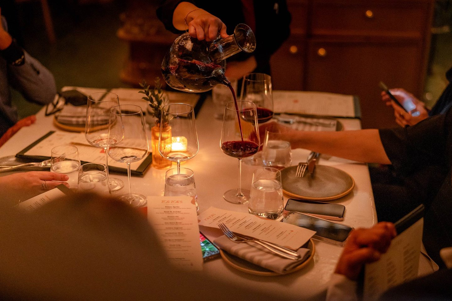 A person pours red wine from a decanter into a glass at a dimly lit restaurant table set with menus, glasses, plates, cutlery, and a candle. Other diners are seated, one holding a phone.