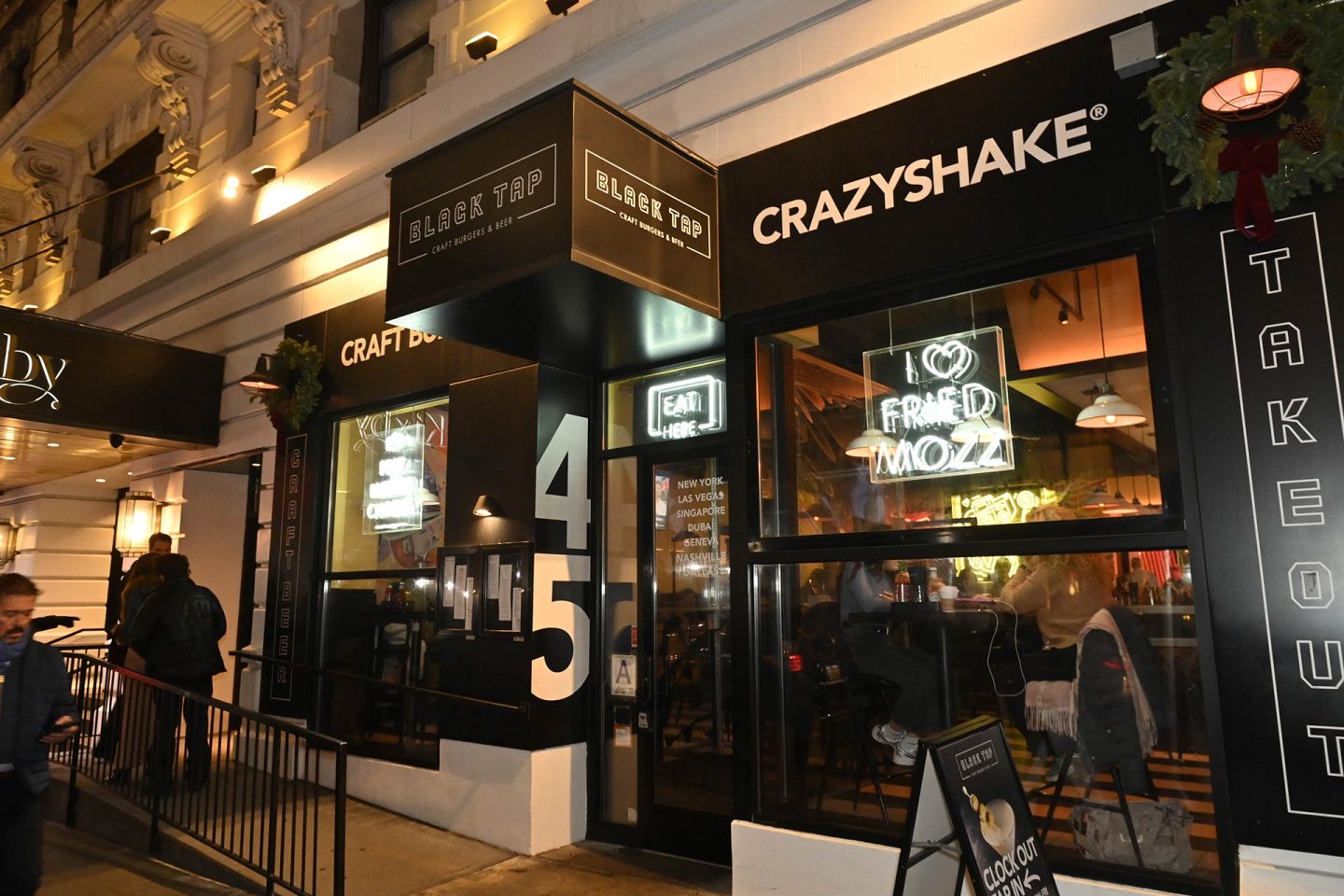 The exterior of Black Tap, a restaurant with black signage and neon lights, including one that says "I ❤️ FRIED MOZZ." People are visible inside, and a menu board stands outside the entrance.