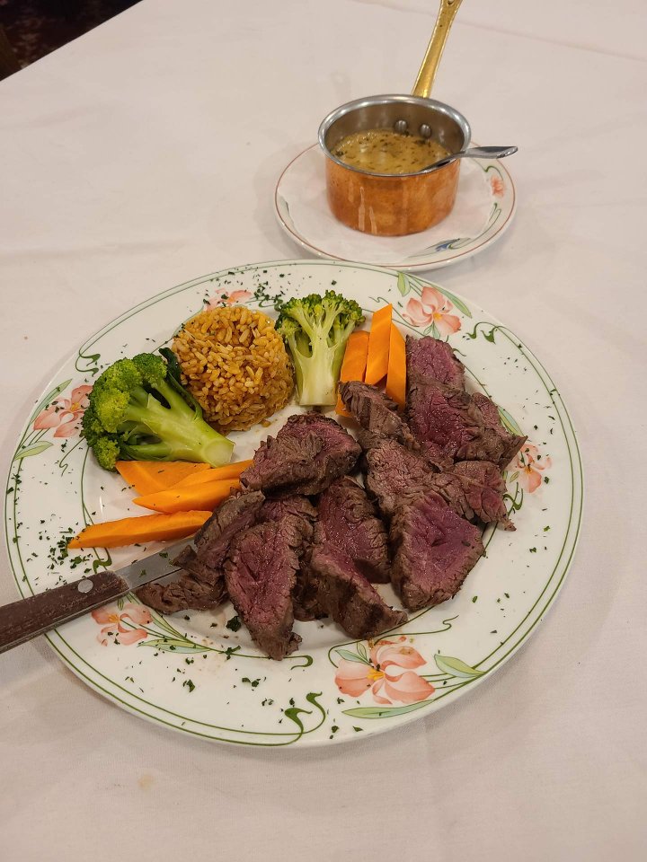 A plate with sliced steak, broccoli, carrot sticks, and a mound of seasoned rice, garnished with herbs. A small pot of creamy sauce sits on a separate plate in the background.