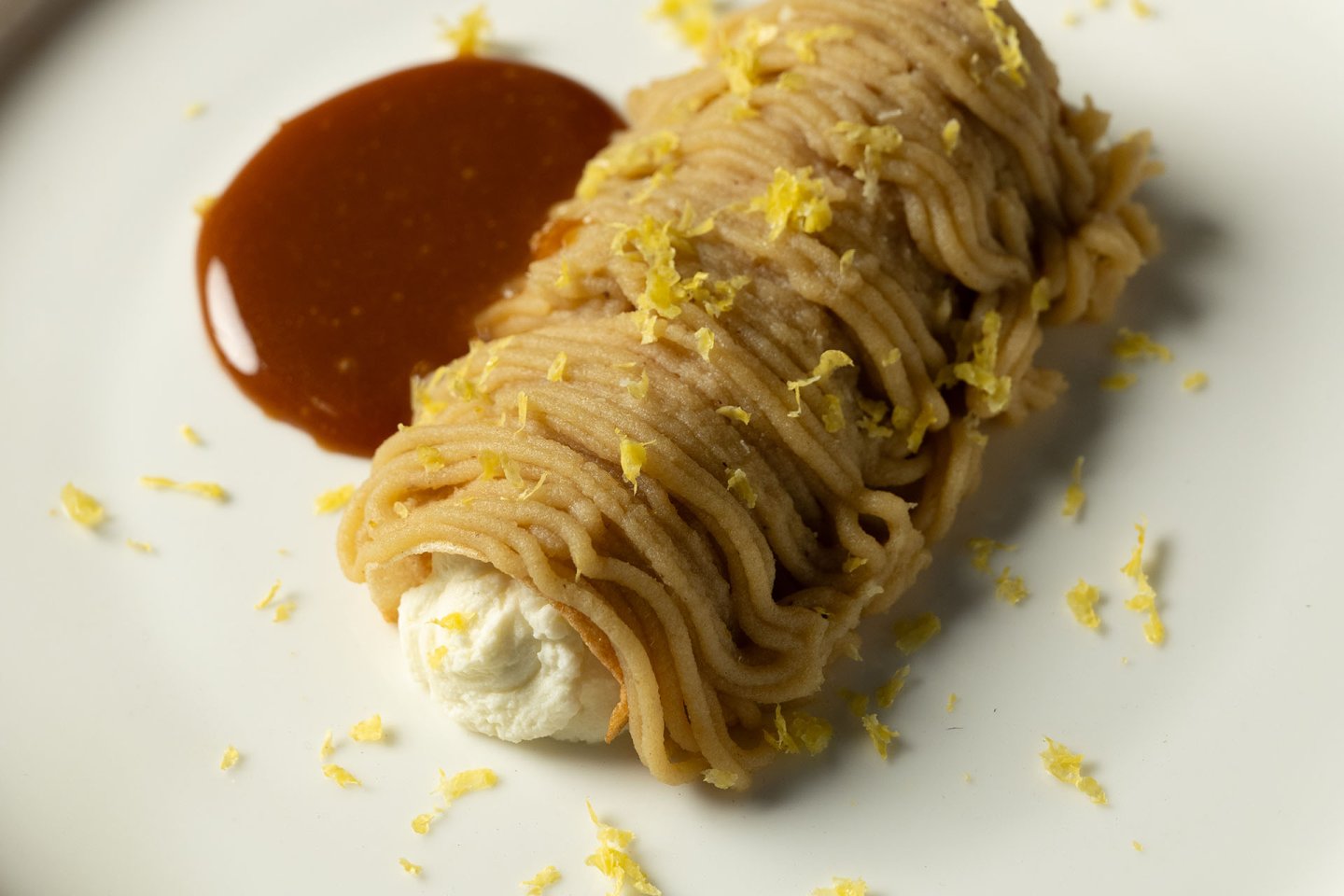 A dessert with piped chestnut cream wrapped around whipped cream, served with a pool of caramel sauce and sprinkled with lemon zest on a white plate.
