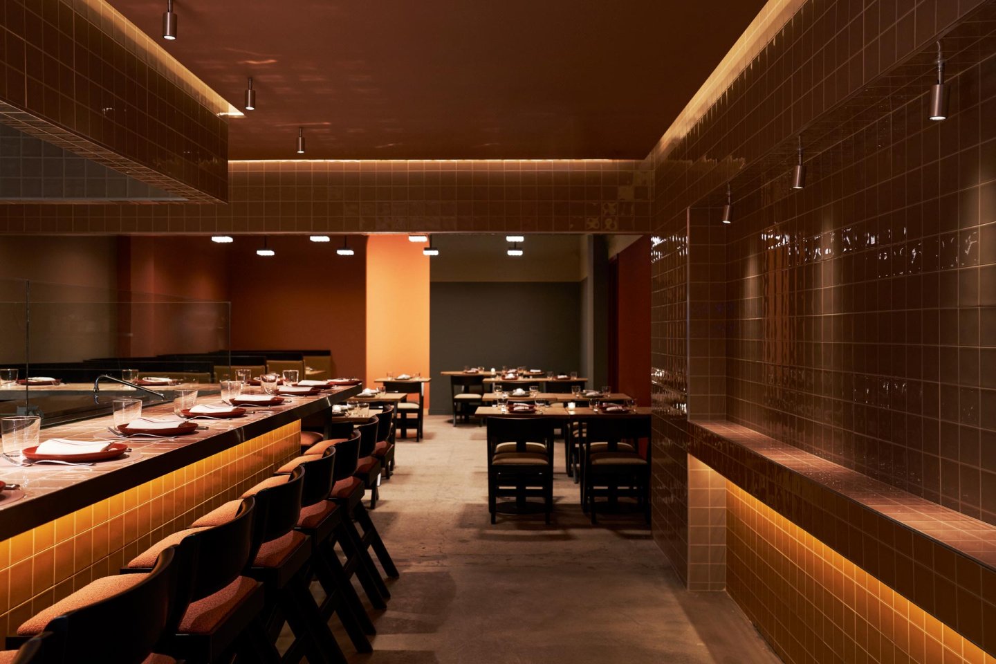 A dimly lit restaurant interior featuring a bar with neatly arranged plates and chairs. The walls and bar are covered in brown tiles, and several tables with chairs are set up in the background. The ambiance is warm and inviting.