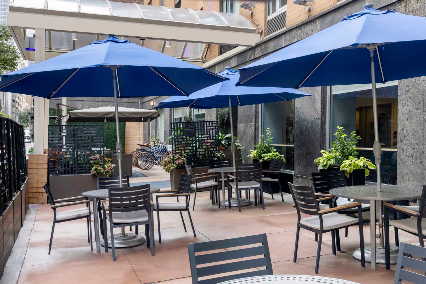 Outdoor dining area with several tables and chairs under blue umbrellas. Potted plants decorate the space, which is enclosed by a modern fence. A bicycle rack and building exterior are visible in the background.