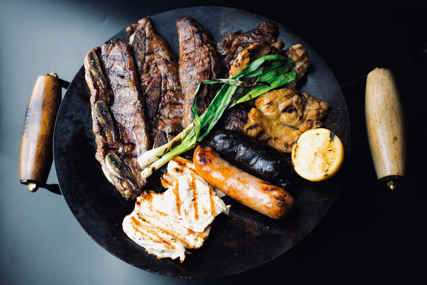 A black round grill pan with wooden handles holds grilled meats, sausages, green onions, a lemon half, and a slice of white cheese, all arranged on a dark background.