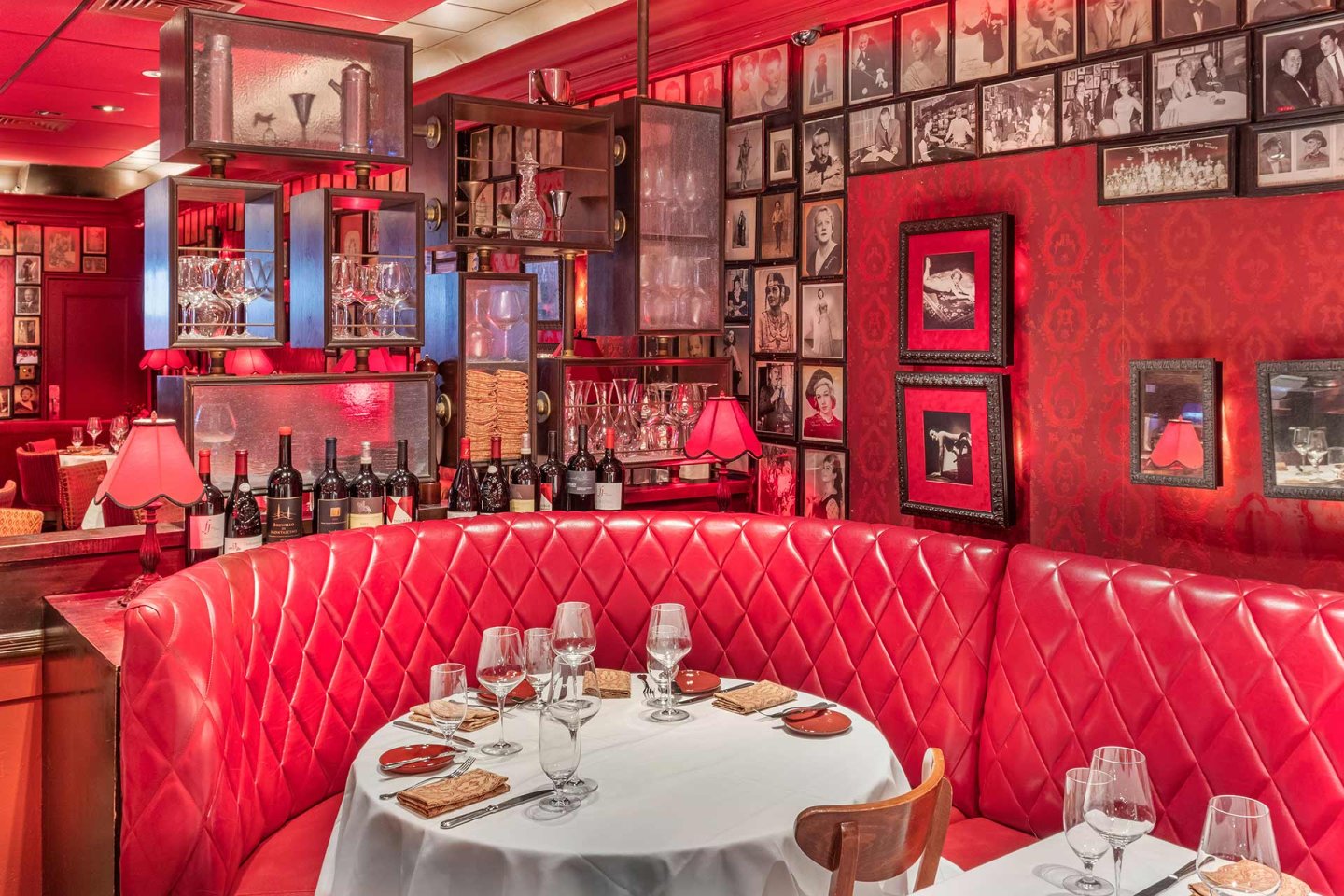 A cozy restaurant features a red leather tufted booth, a round table set with wine glasses and plates, warmly lit by red lamps, with walls covered in framed black-and-white photos and patterned red wallpaper.