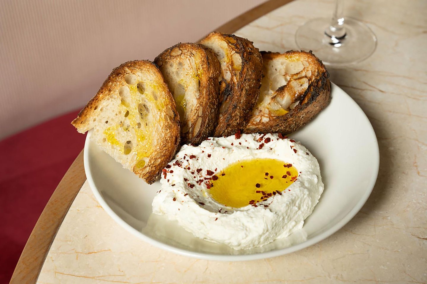 Four slices of toasted bread are served on a plate with whipped ricotta cheese, topped with olive oil and red pepper flakes. A wine glass is partially visible on the marble table.