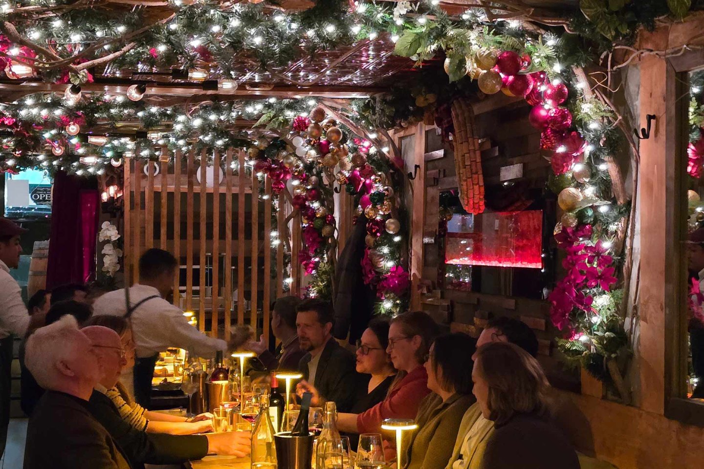 A group of people sits around a long table in a warmly lit restaurant decorated with garlands, flowers, and string lights. A waiter attends to them. The atmosphere is festive and cozy.