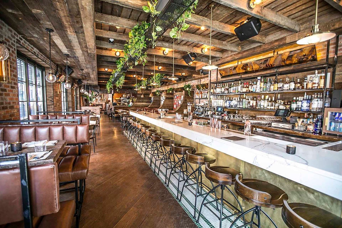 A rustic bar interior with a long white countertop, surrounded by metal stools. Wooden beams and plant decorations hang from the ceiling. Shelves stocked with bottles line the brick walls. Leather booths occupy the left side. Warm ambient lighting.