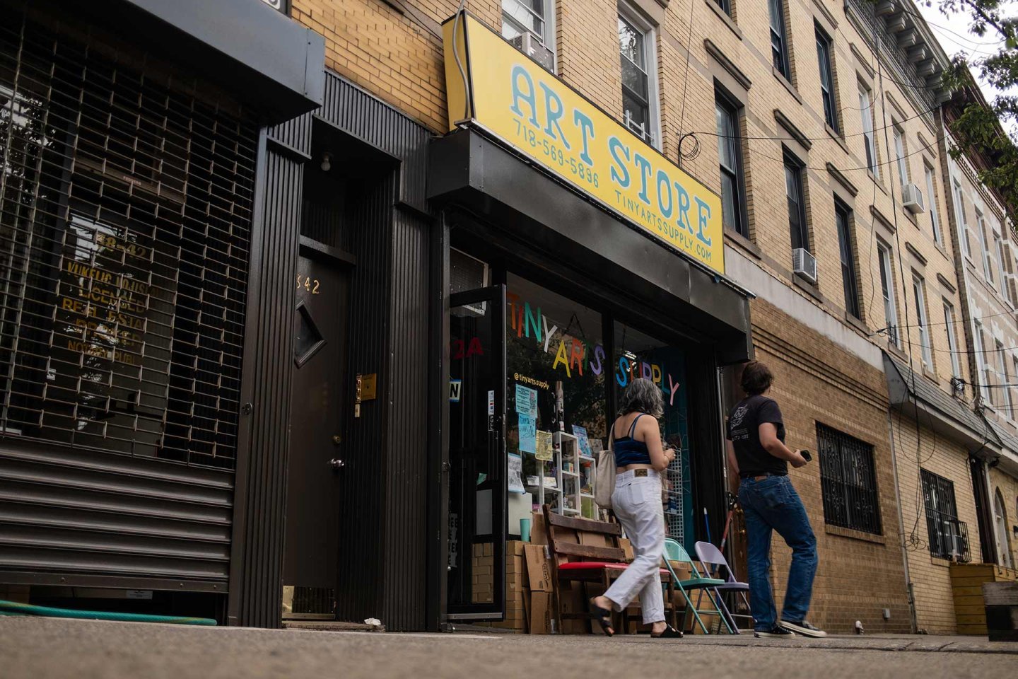 Two people walk past a storefront with a yellow “ART STORE” sign. The shop windows display various art supplies and colorful decorations. The building exterior is tan brick, and the street appears quiet.