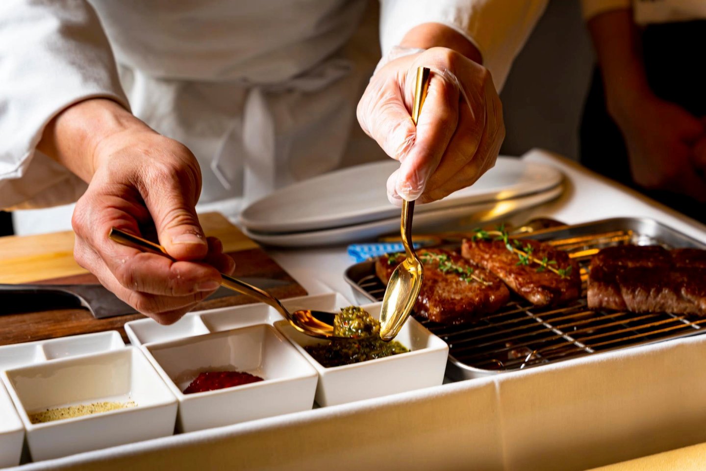 A chef's hands preparing a dish, with one hand holding a spoon over a container with herbs. Grilled meat rests on a rack beside several small bowls of spices and herbs. Plates are stacked in the background.