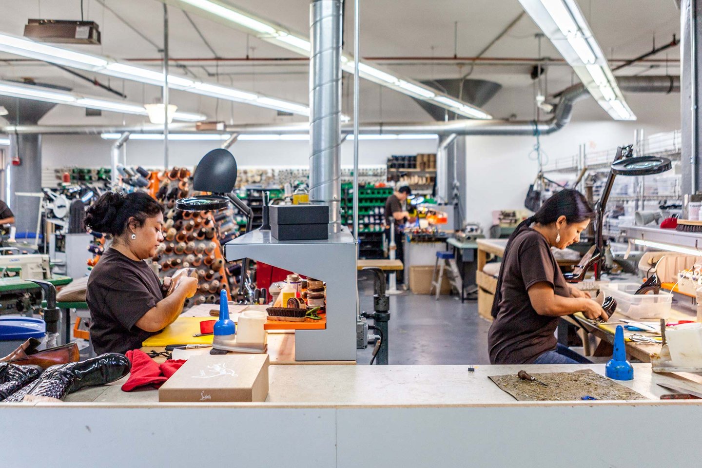 Two women work at separate stations in a well-lit factory, using tools and machinery to craft or repair leather goods. Shelves with materials and equipment are visible in the background.