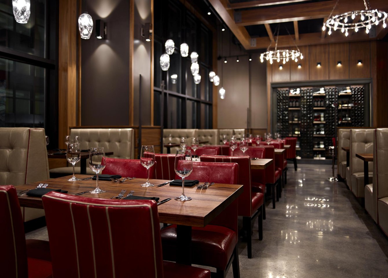 Modern restaurant interior with red leather chairs, wooden tables set with wine glasses and silverware, beige upholstered booths, wine rack, large windows, and decorative pendant lights. The space is empty and well-lit.