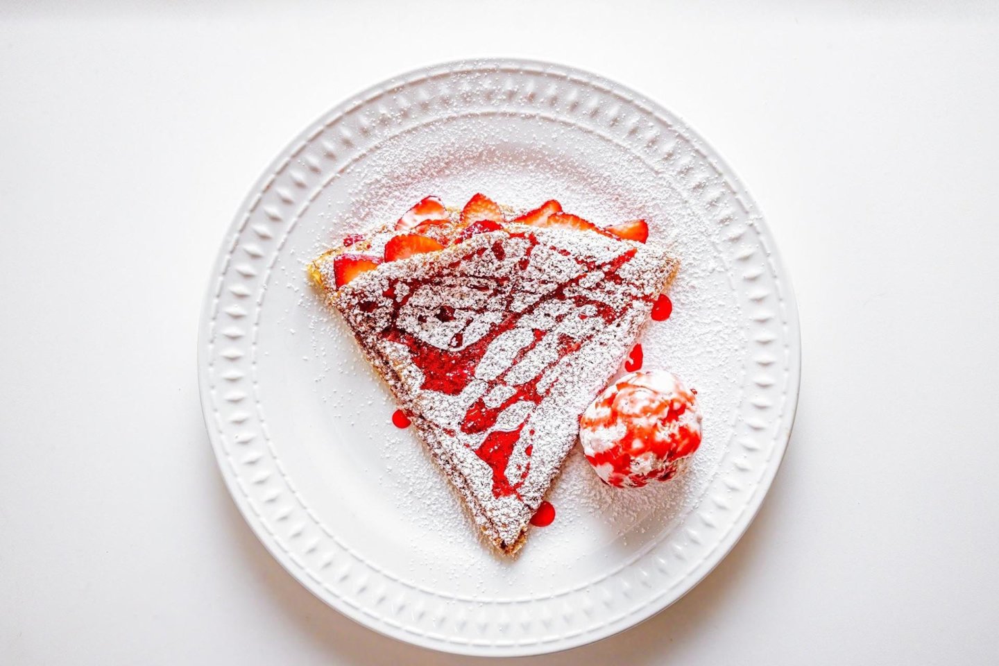 A white plate with a triangle of French toast topped with sliced strawberries, powdered sugar, and strawberry sauce, served with a scoop of ice cream, on a white background.