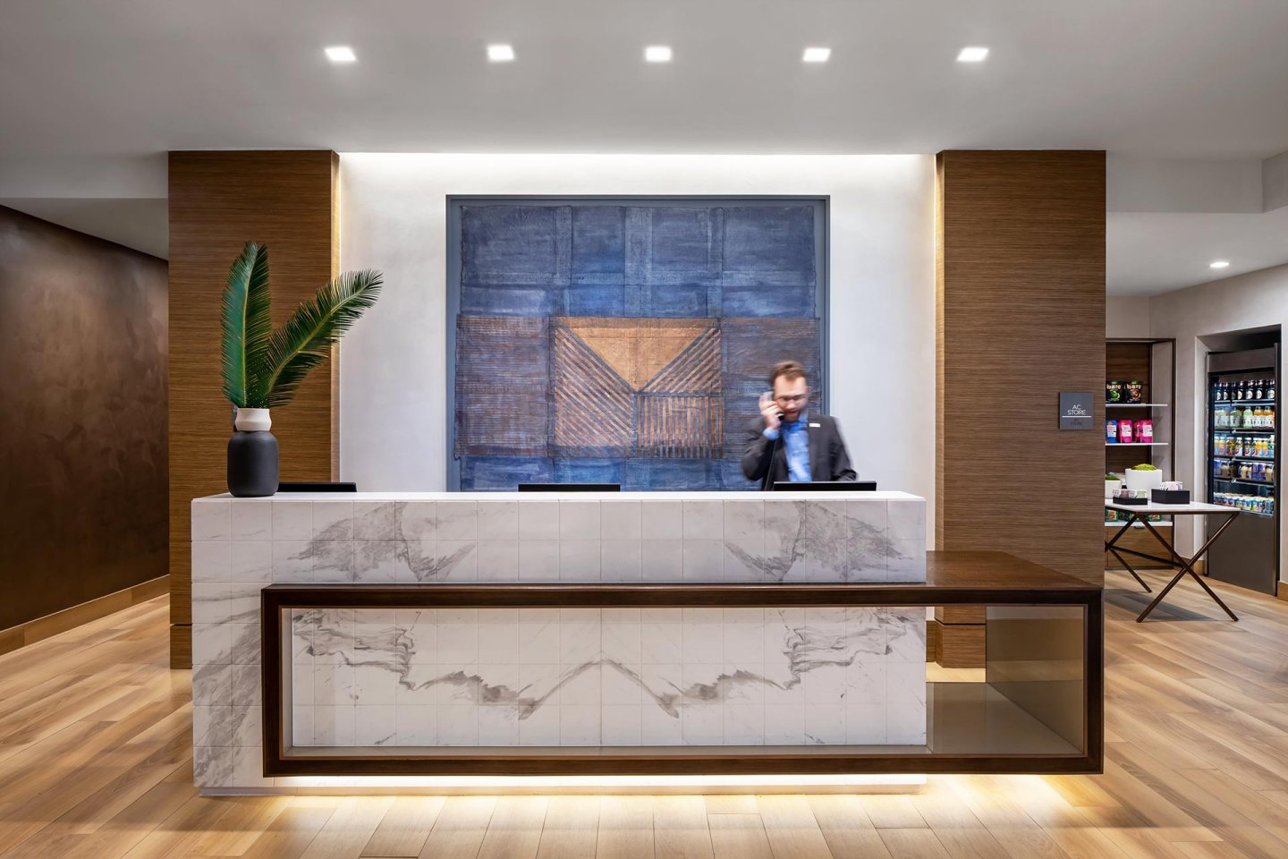 A modern hotel reception desk with a marble finish, a large blue abstract painting behind it, and a potted plant on the counter. A man in a suit stands behind the desk, talking on the phone. Shelves with snacks are visible to the right.