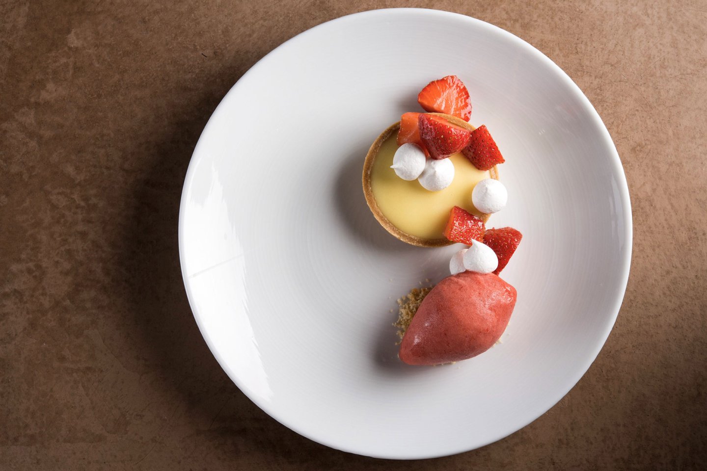 A white plate with a lemon tart topped with strawberries and small meringues, next to a scoop of strawberry sorbet, set on a brown textured surface.