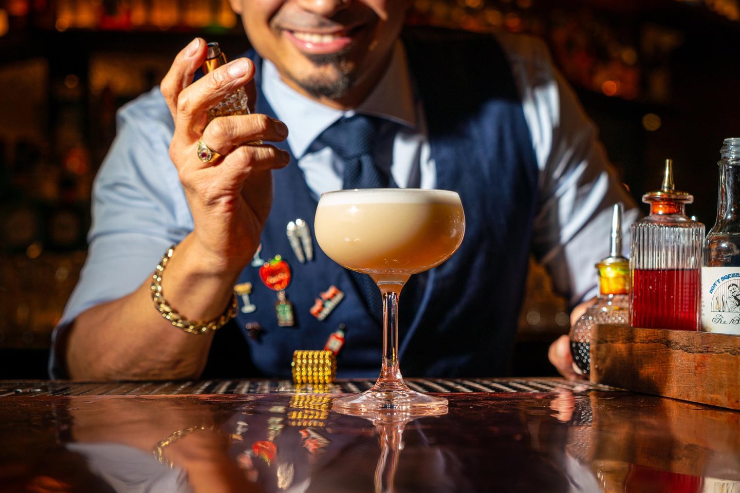 A bartender in a vest and tie sprays garnish over a frothy cocktail in a coupe glass, with various bar tools and bottles on the counter. The scene is warmly lit, capturing a joyful expression.