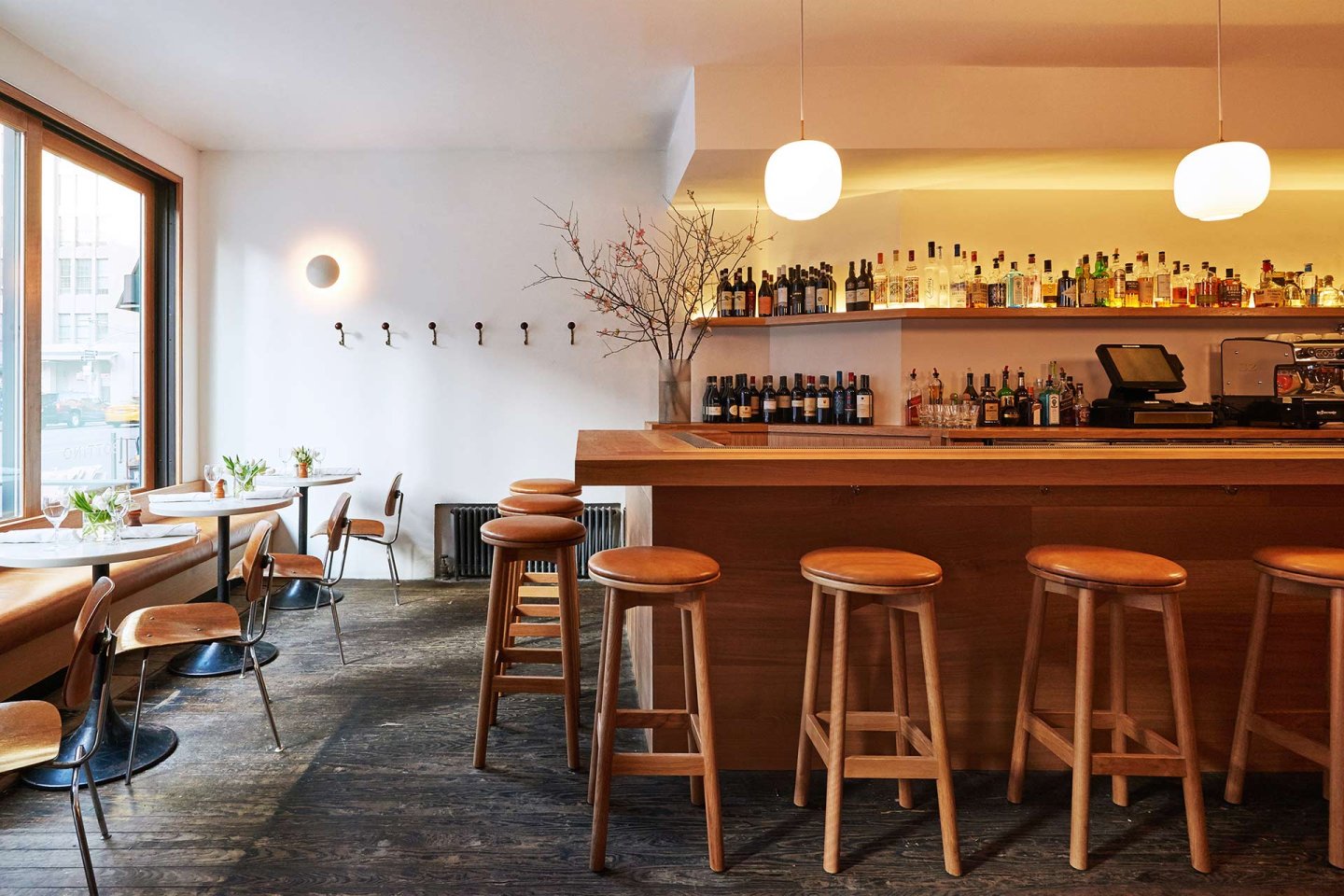A modern bar with wooden stools, a long counter, shelves stocked with bottles, and small tables with chairs by large windows. Minimalist decor, white walls, and warm lighting create a cozy, inviting atmosphere.