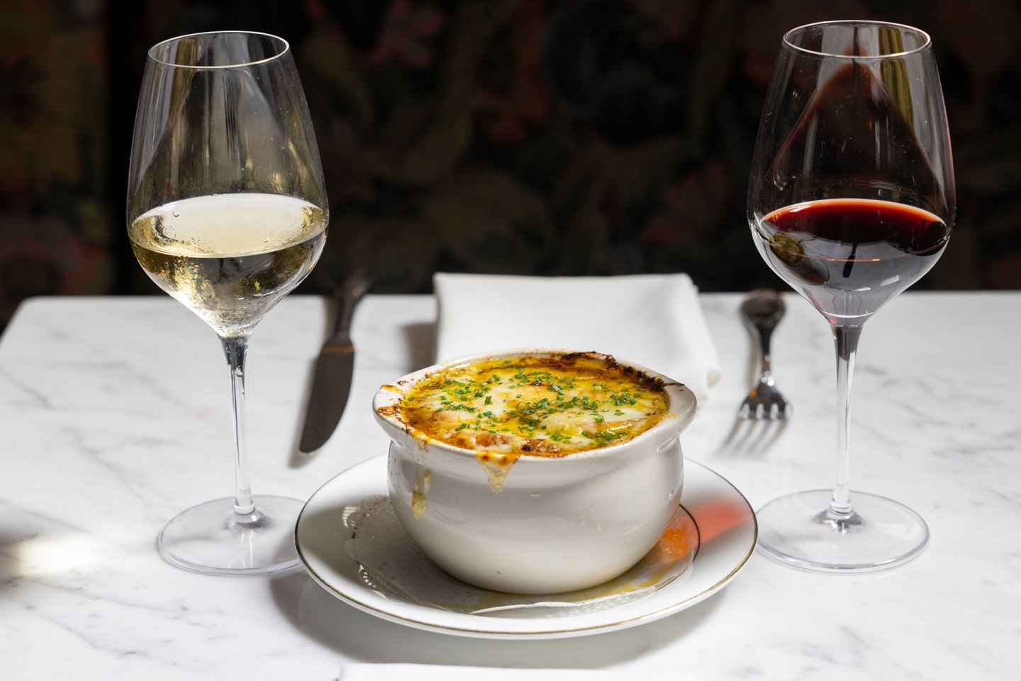 A bowl of French onion soup topped with melted cheese sits on a white plate, flanked by glasses of white and red wine on a marble table, with silverware and a white napkin in the background.