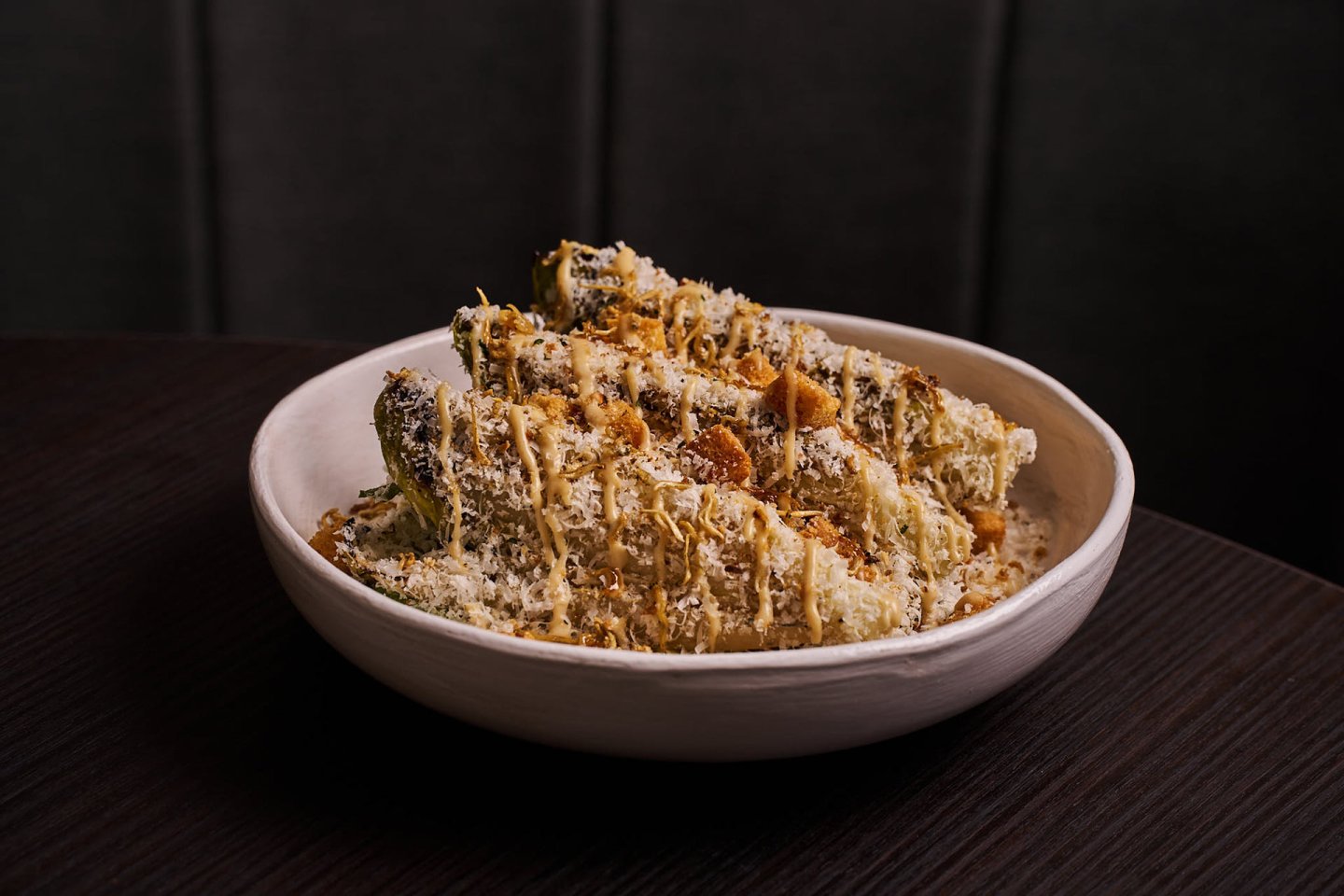 A white bowl filled with breaded, baked zucchini fries topped with grated cheese and seasoning, placed on a dark wooden table.