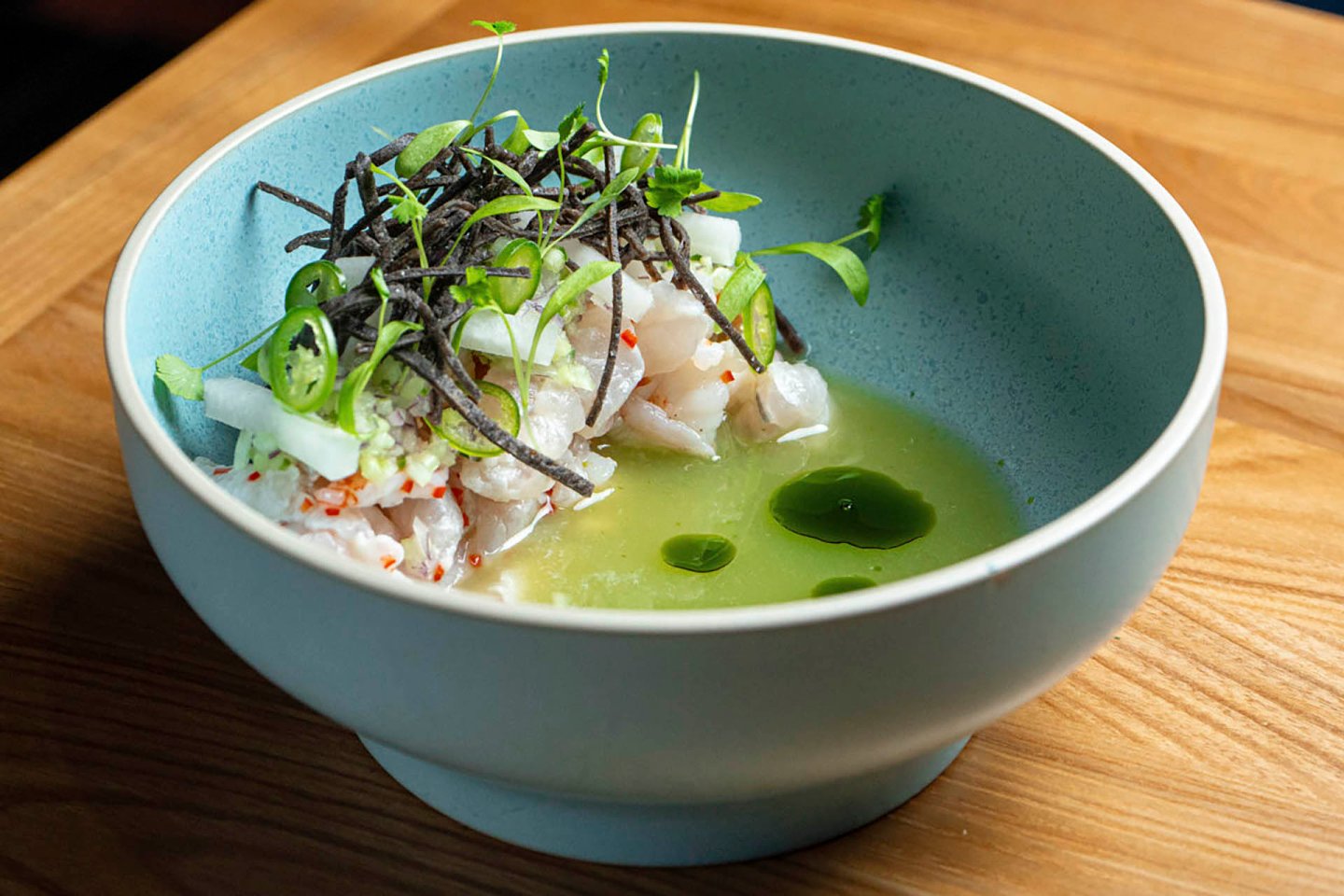 A light blue bowl containing diced raw fish dressed in a green sauce, garnished with thin black strips and microgreens, sits on a wooden table. The dish is artfully presented, highlighting its fresh ingredients.