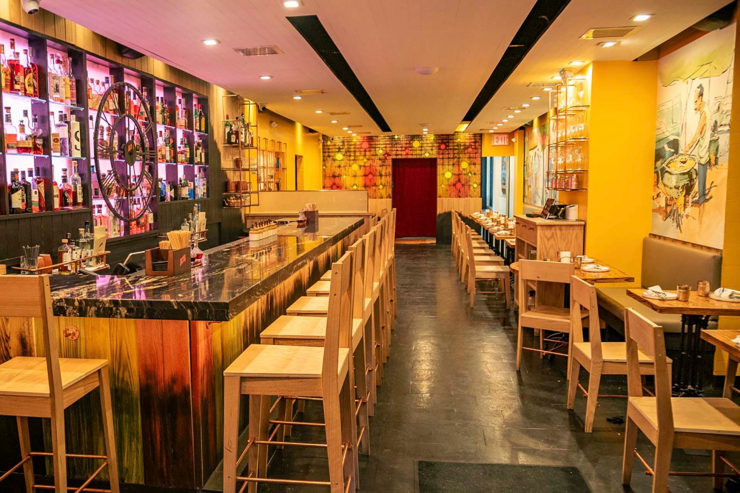A modern bar and restaurant with a long marble-topped bar, high wooden chairs, and shelves displaying various bottles. The interior features warm lighting, yellow walls, artistic murals, and neatly arranged tables along the opposite side.