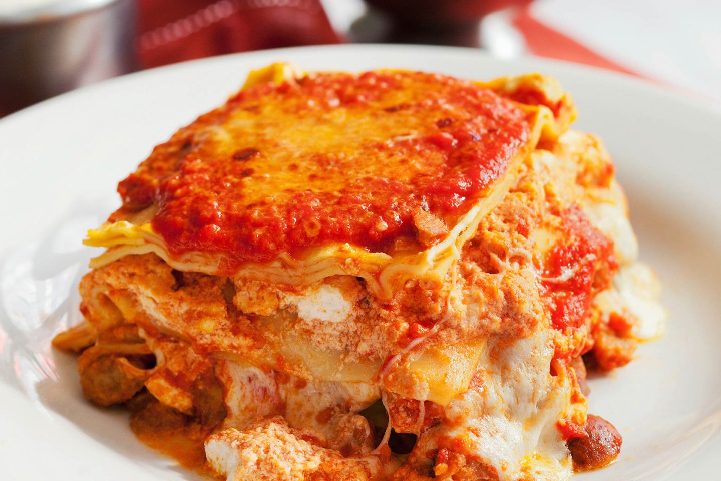 A close-up of a slice of lasagna on a white plate, showing layers of pasta, melted cheese, meat, and rich tomato sauce.