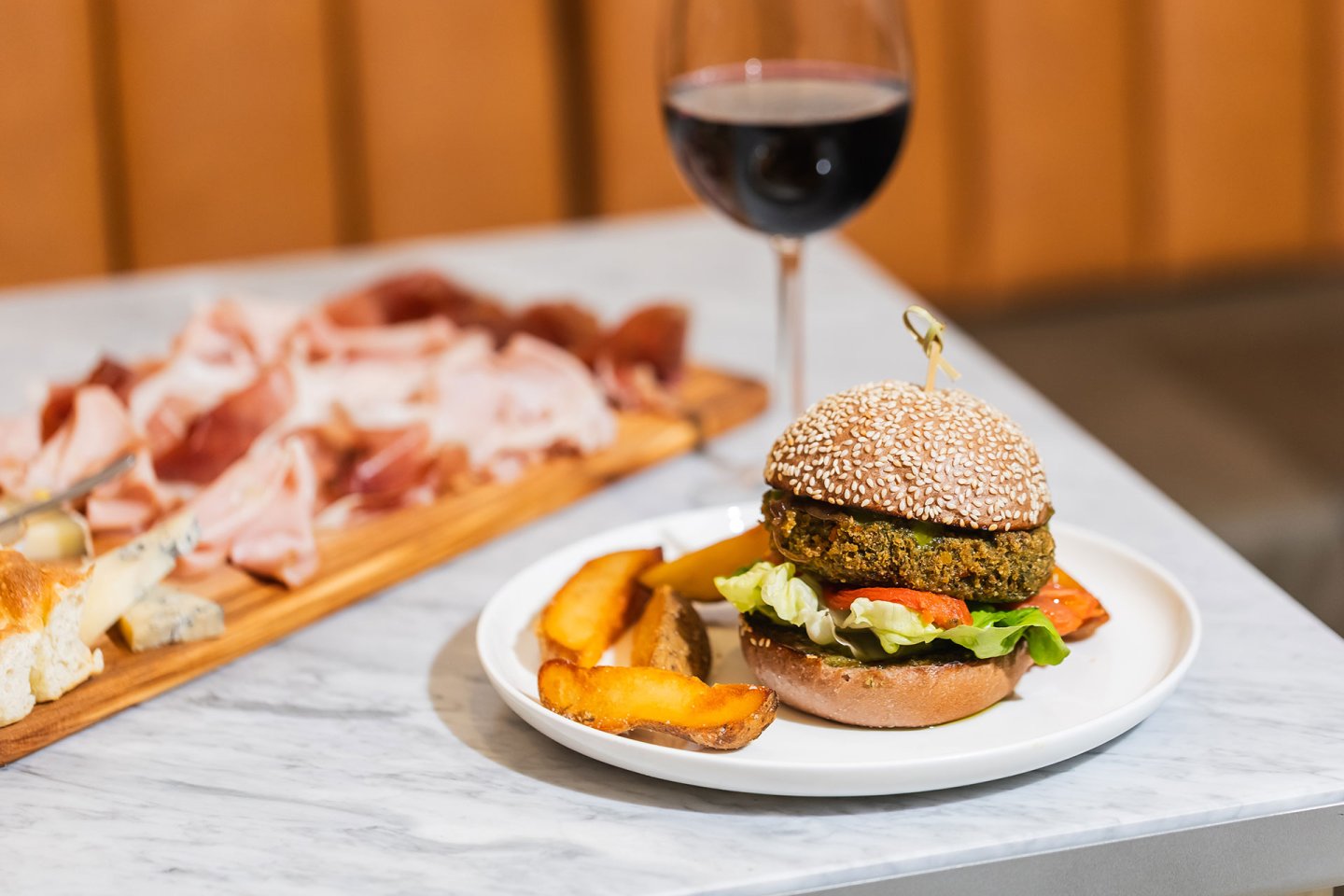 A veggie burger with lettuce and tomato, served with potato wedges on a white plate, sits on a marble table next to a glass of red wine and a wooden board with assorted sliced meats and cheeses.