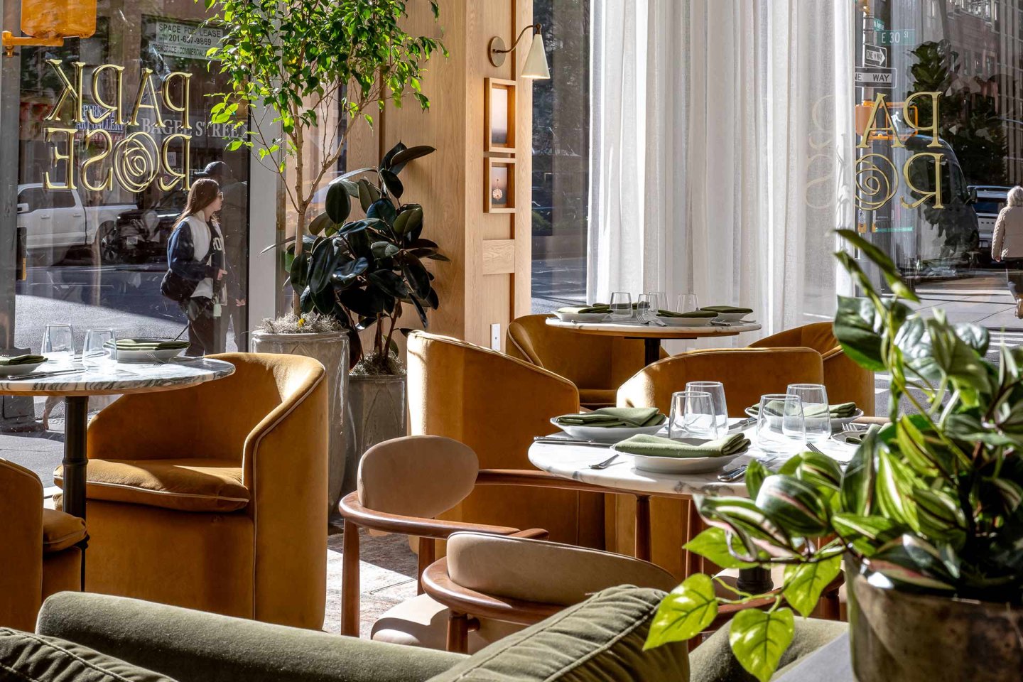 A sunlit restaurant interior with tan chairs, green plants, set tables, and large windows showing a street scene outside. Light streams in, creating a bright, inviting atmosphere.