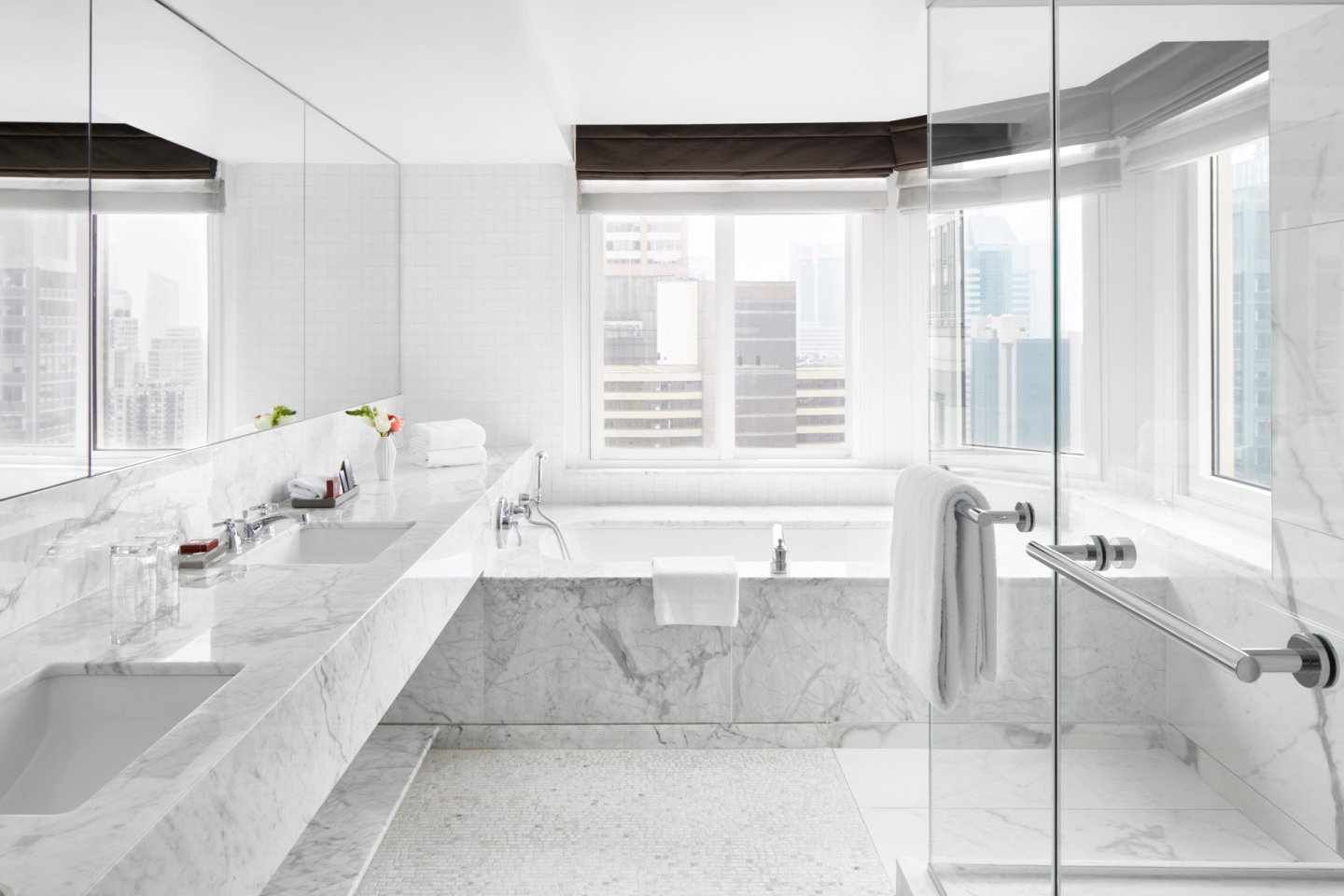 Modern bathroom with marble countertops, double sinks, a glass shower, and a bathtub by large windows overlooking a cityscape. White walls and natural light create a bright, spacious atmosphere.