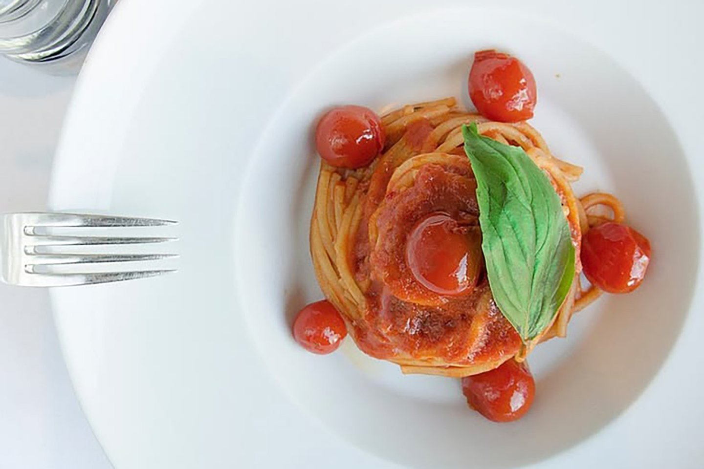 A plate of spaghetti topped with tomato sauce, cherry tomatoes, and a fresh basil leaf, with a fork and a glass at the edge of the white plate.