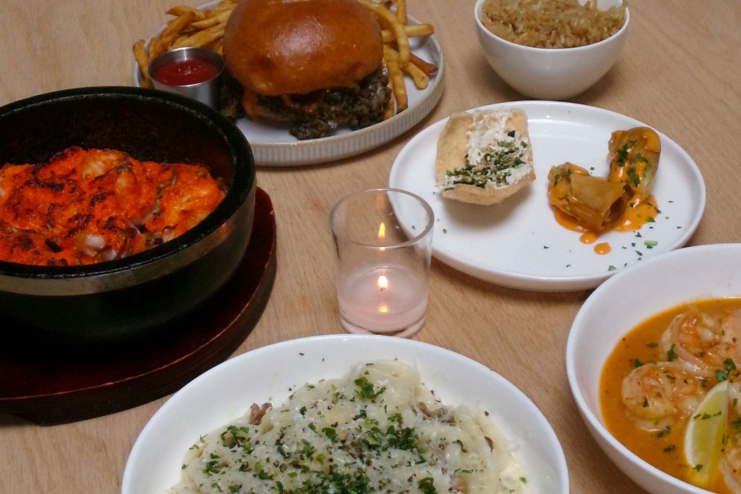 A table with various dishes including pasta, shrimp, dumplings, bread with spread, rice, a burger with fries, and a candle in the center.