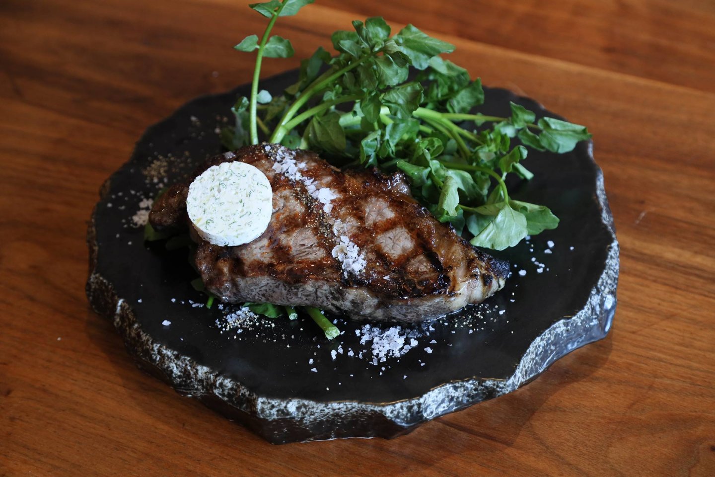 A grilled steak with grill marks, topped with a pat of herb butter and sprinkled with coarse salt, is served on a rustic black stone plate with fresh green garnish.