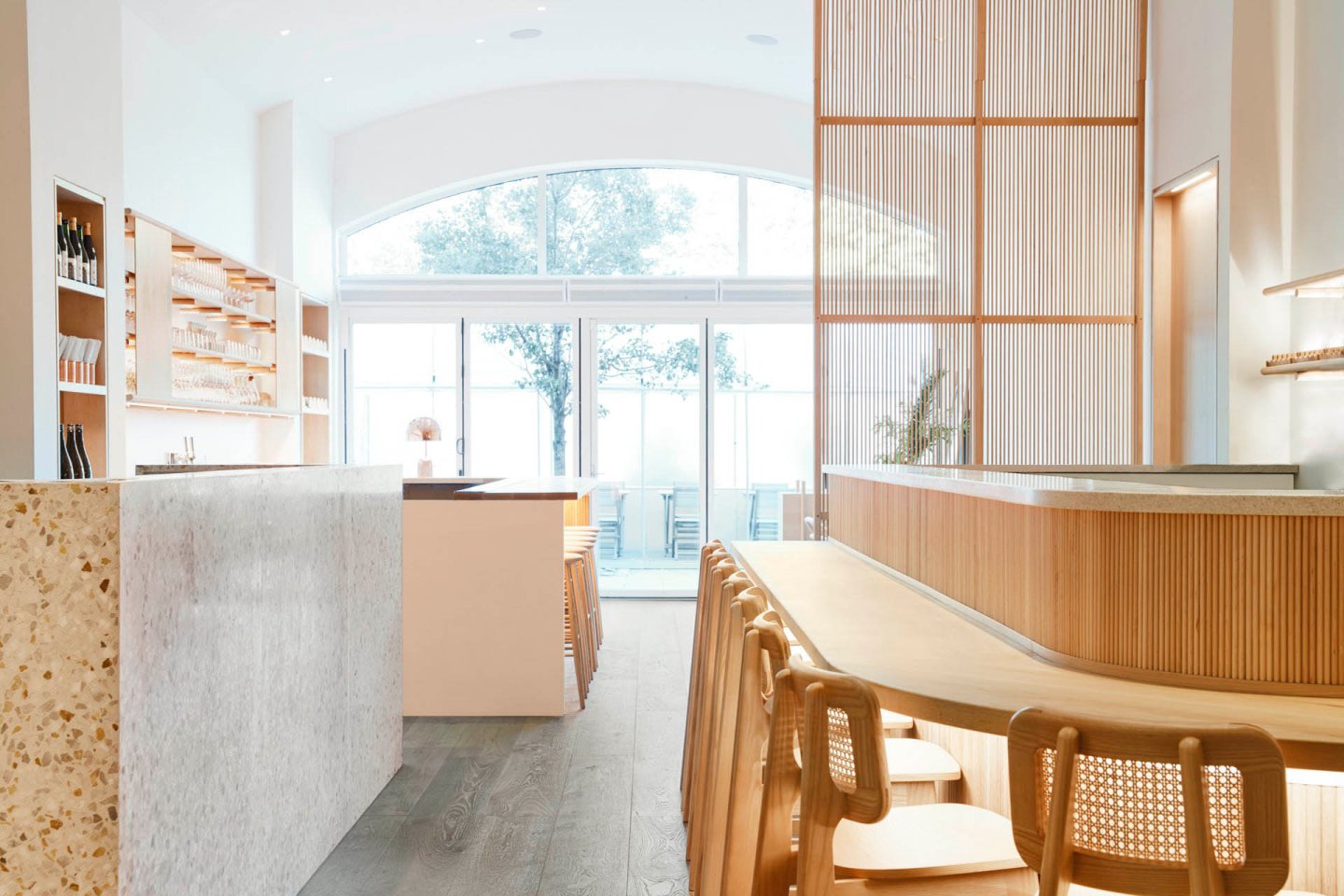 Bright, minimalist cafe interior featuring a large window, light wooden furniture, and stone counters. Rows of chairs line the counters, and shelves with bottles and items are on the left wall. Light streams in, creating an airy atmosphere.