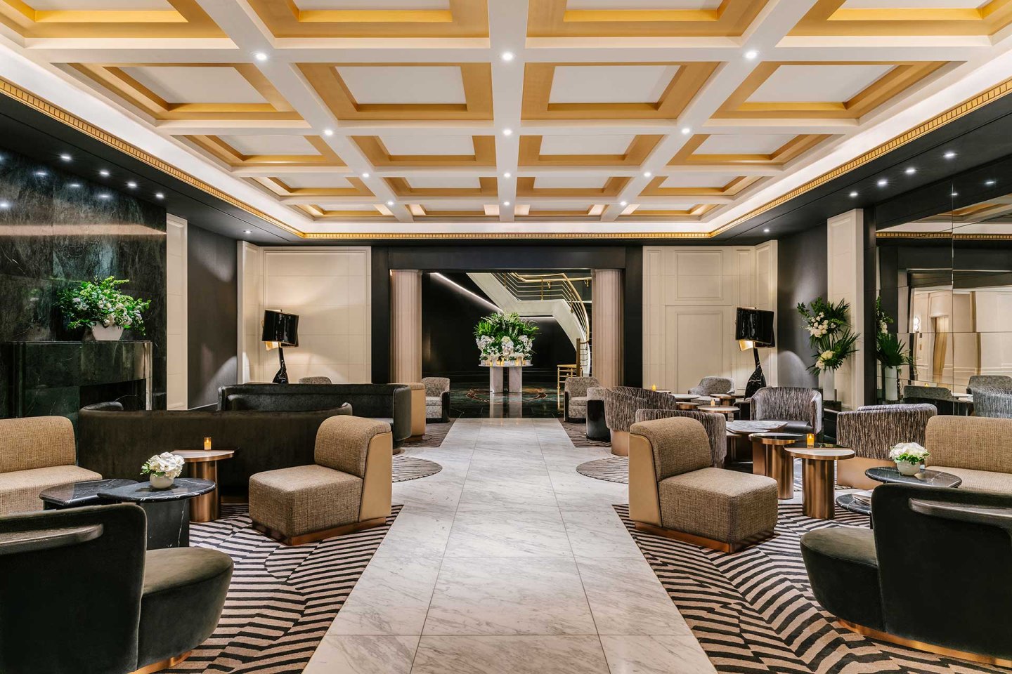 Luxurious hotel lobby featuring modern armchairs, sofas, and low tables on patterned carpets, with marble flooring, decorative ceiling panels, soft lighting, and plants adding a touch of greenery.
