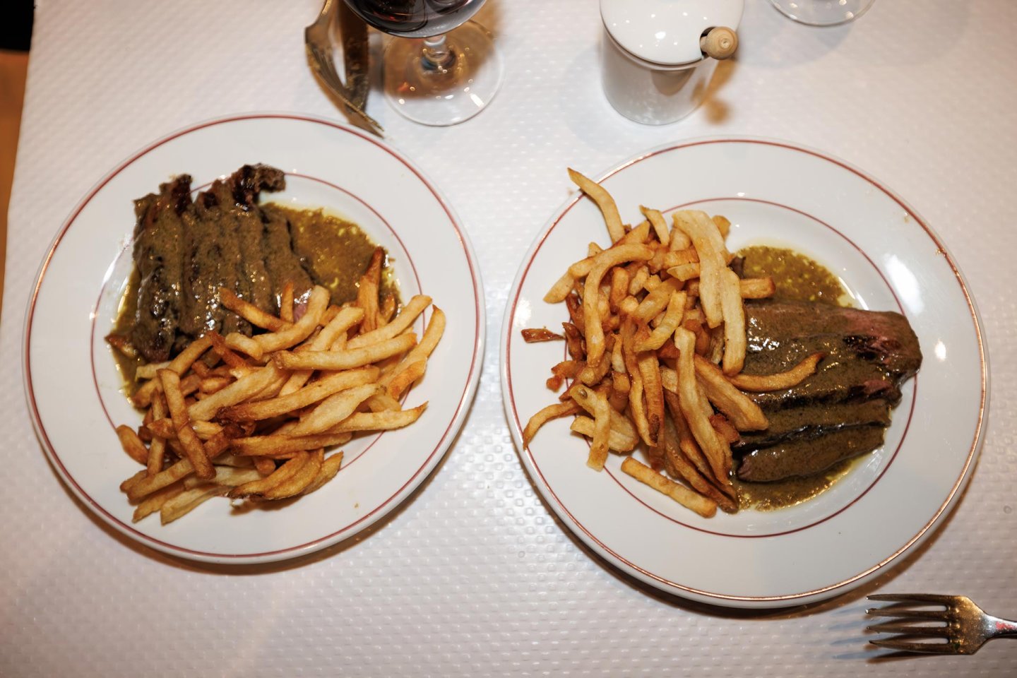 Two plates of steak with brown sauce and French fries on a white tablecloth, viewed from above. A glass of red wine, a small white pot, and part of a fork are also visible on the table.