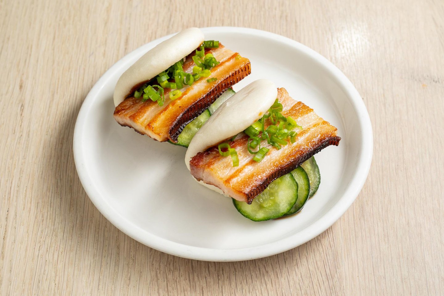 Two steamed buns filled with crispy pork belly, cucumber slices, and chopped green onions are served on a white plate atop a light wooden table.