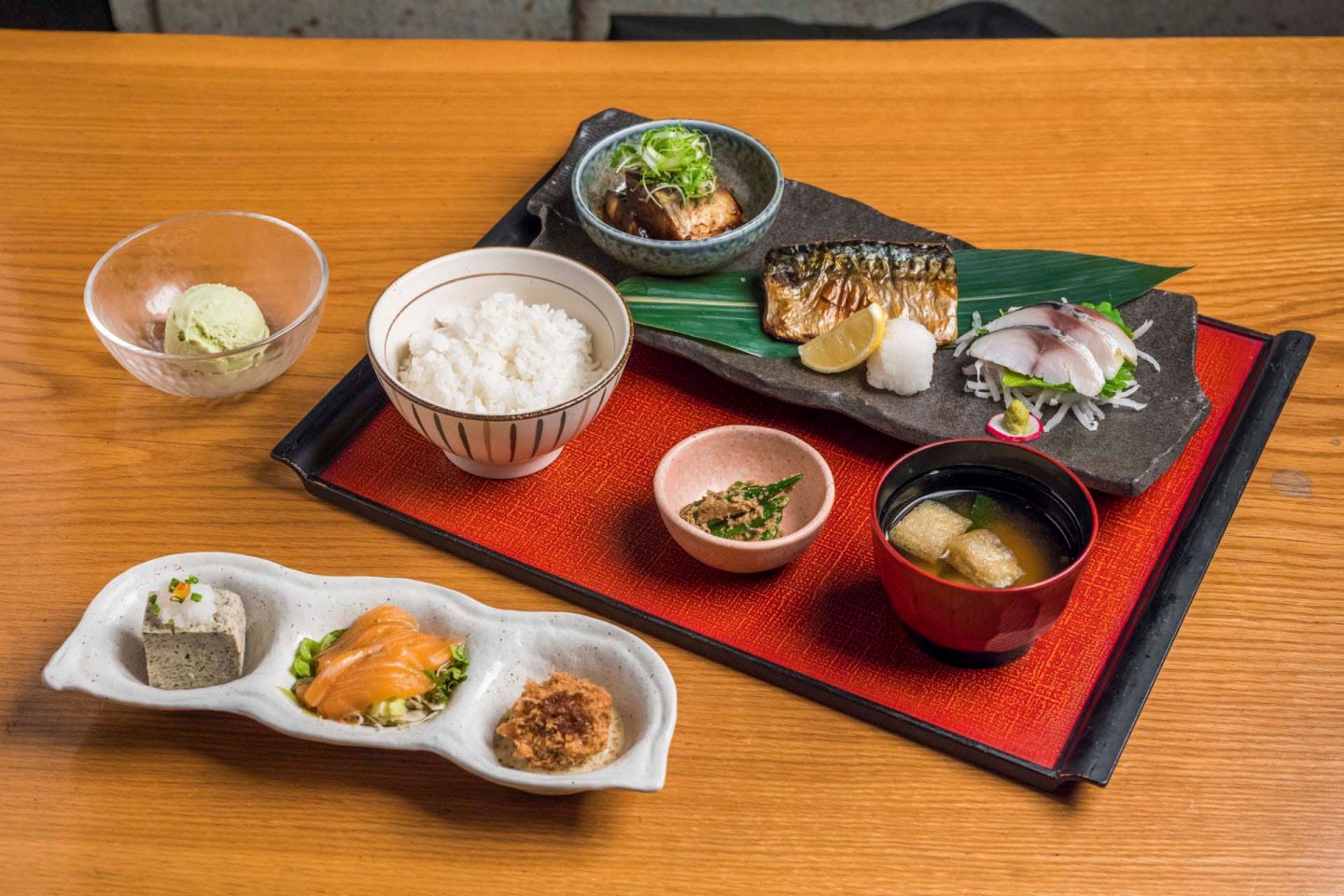 A Japanese meal set on a wooden table. It includes grilled fish, white rice, miso soup, pickles, tofu, and a dessert with green ice cream. The dishes are elegantly arranged on different plates and bowls.