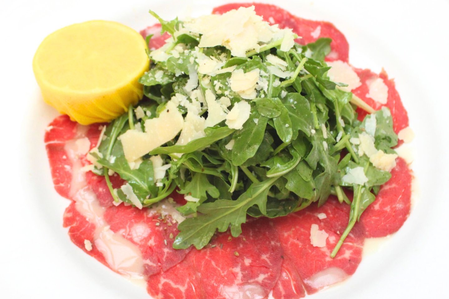 Thinly sliced raw beef topped with arugula and shaved Parmesan on a white plate. A lemon half sits to the side, enhancing the presentation of this classic carpaccio dish.