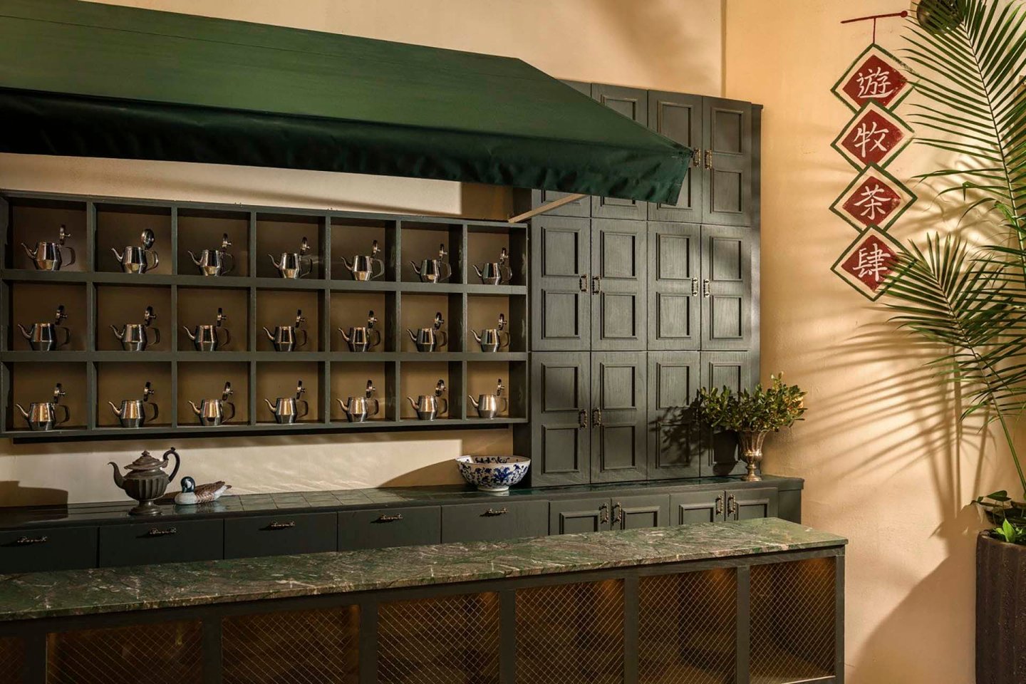 A serene tea shop features dark green cabinetry with multiple shelves displaying teapots. A green awning provides shade, and red Chinese decorations hang on the wall. A palm plant adds greenery to the setting, creating a peaceful atmosphere.