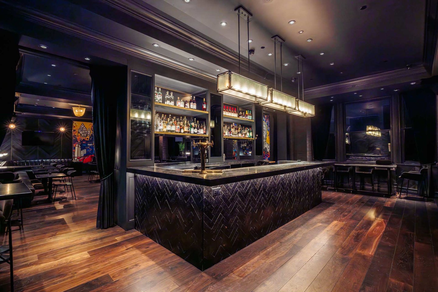 A modern bar with dark wood floors, black herringbone-patterned bar, illuminated shelves stocked with bottles, pendant lighting, and black barstools in a dim, stylish interior.