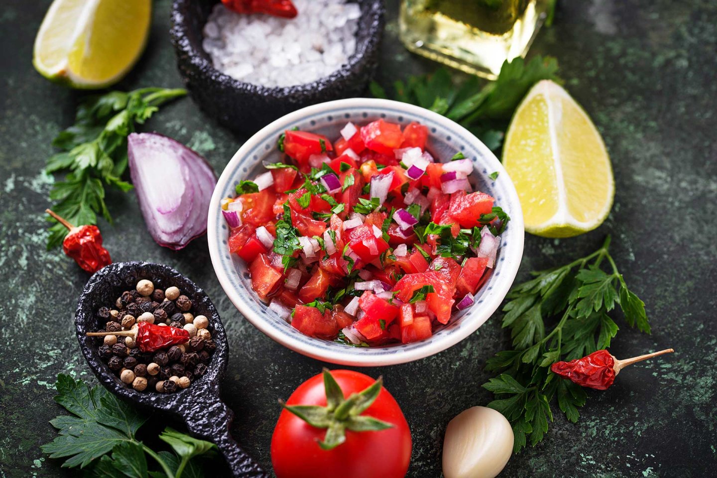 A bowl of fresh salsa with diced tomatoes, onions, and cilantro, surrounded by lime wedges, parsley, a tomato, garlic, spices, and coarse salt on a dark surface.