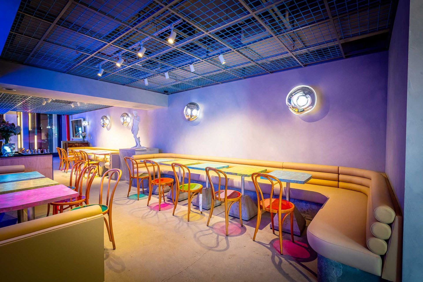 A stylish modern cafe with pastel walls, wooden chairs, colorful table tops, and soft lighting from round wall fixtures. A cushioned bench runs along the wall, creating a cozy, inviting atmosphere.