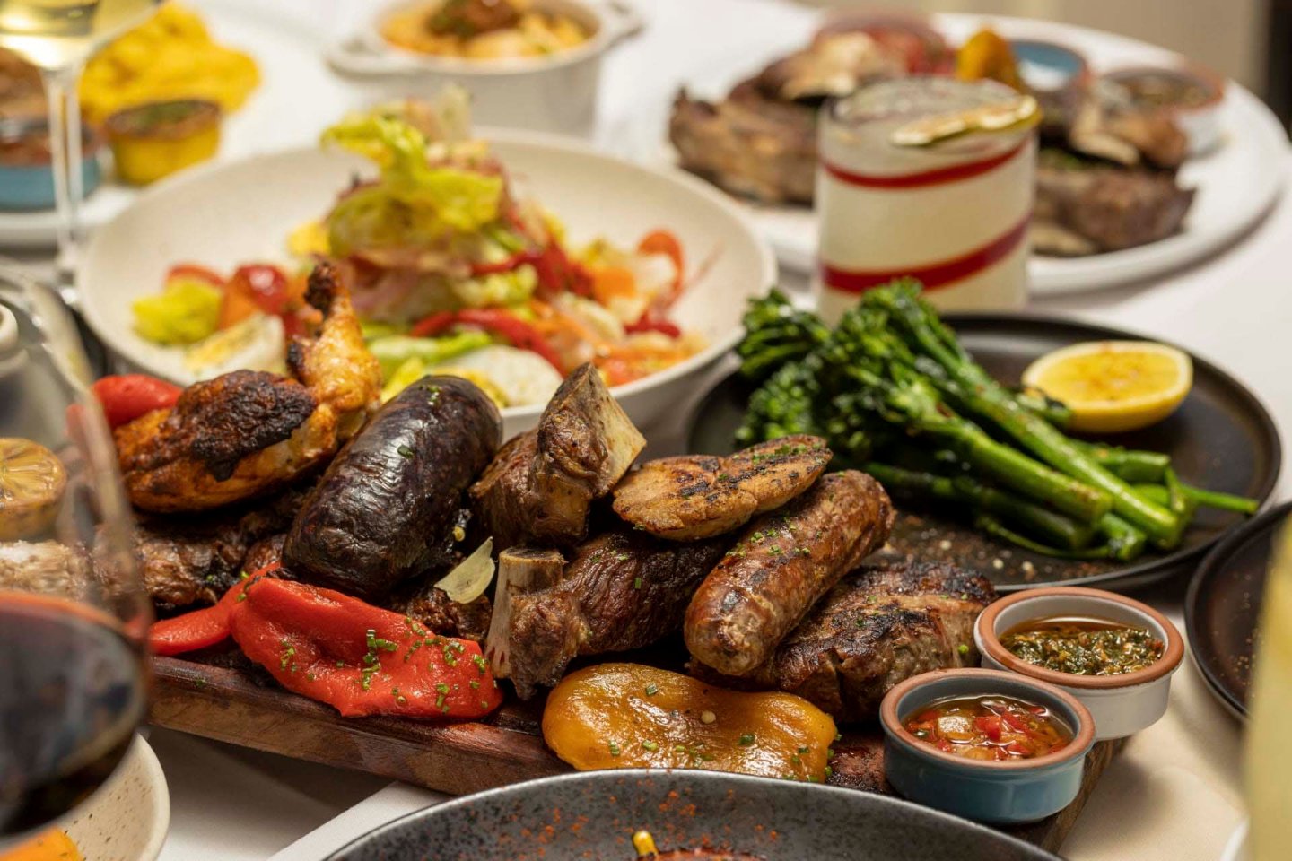 A platter of grilled meats including sausages and chicken, alongside roasted vegetables. Accompanied by small dishes of sauces and a salad. A side of asparagus and lemon, with wine glasses and more dishes in the background.