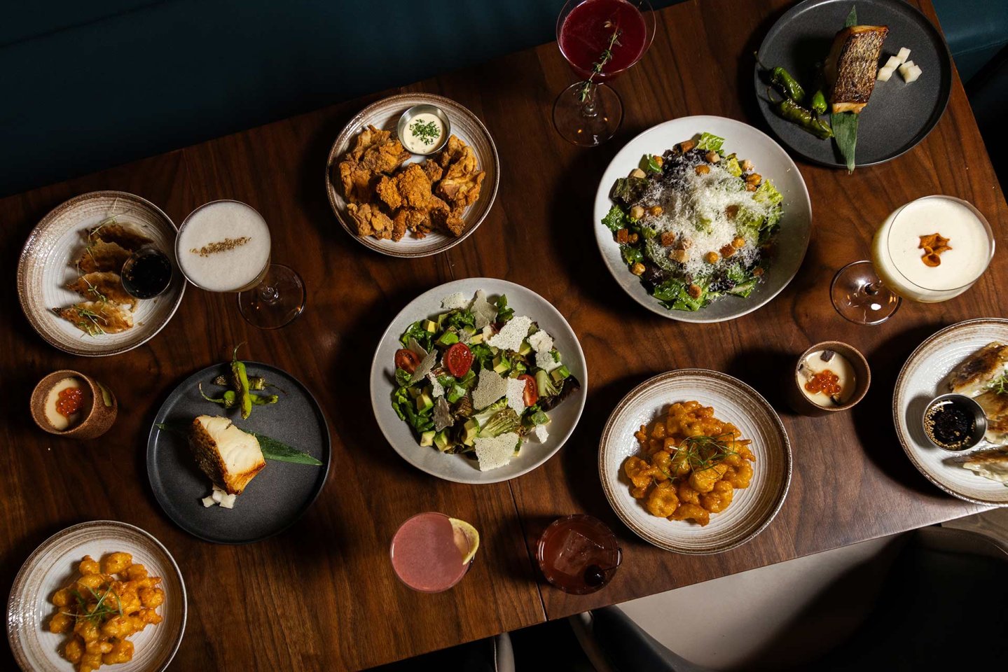 An overhead view of a wooden table set with assorted dishes, including salads, pasta, fried foods, bread, and various cocktails and desserts, arranged for a group meal.