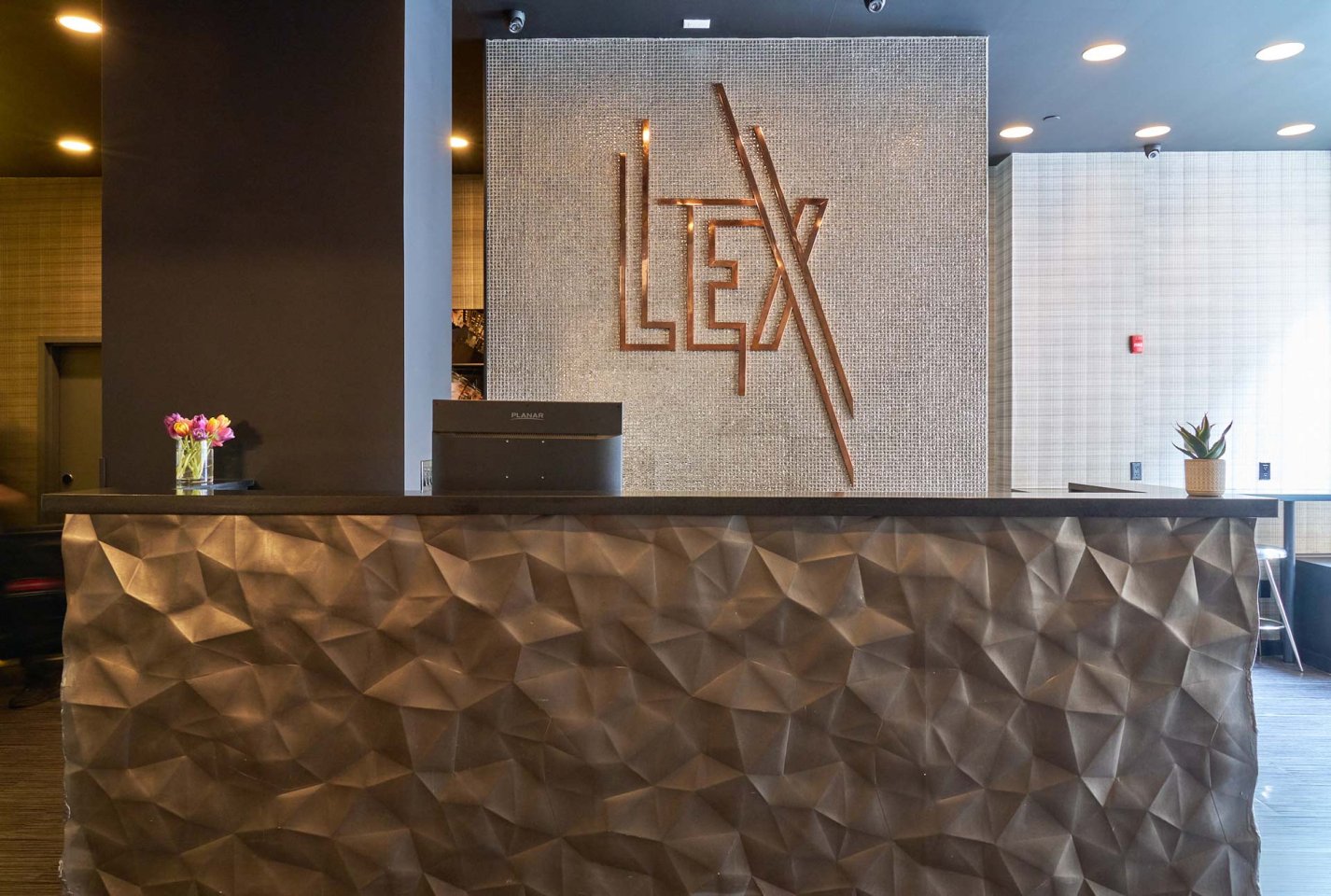 Modern hotel reception desk with a geometric textured front, small flower vase and plant on the counter, and a wall behind displaying large metallic letters spelling “LEX.” Warm lighting creates a welcoming atmosphere.