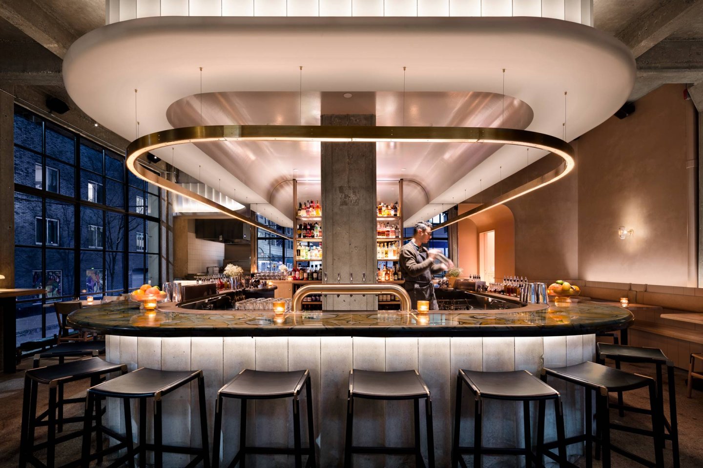 A modern, stylish bar with a curved counter, high stools, soft lighting, and a bartender preparing drinks. Shelves of bottles are illuminated behind the bar, and large windows let in natural light.